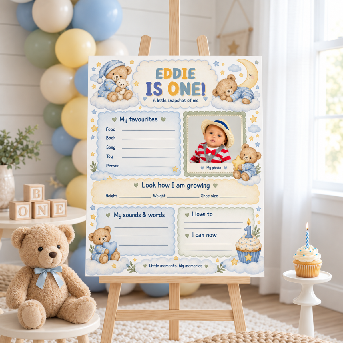 Boy's teddy bear first birthday milestone poster with a photo area and blue memory sections, on an easel beside teddy bears.