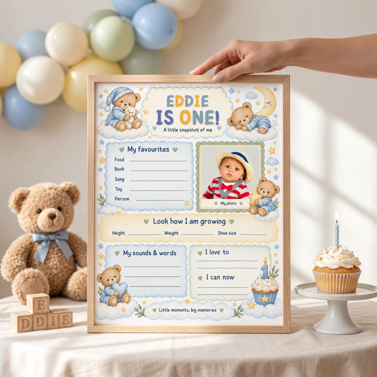 Boy's teddy bear first birthday milestone poster with a photo area and blue memory sections, framed poster held on a table.